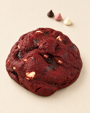 A large red velvet chocolate chip cookie with white chocolate chips, photographed on a light beige background with three small chocolate kisses in the background.